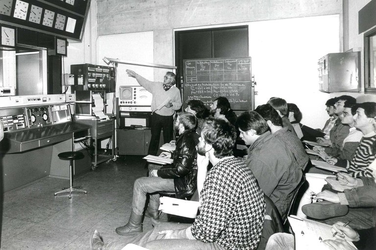 Schwarz-Weiß-Foto: Rechts im Bild sitzt eine GruppeStudenten, links im Bild ein Dozent, der auf die Gerätschaften links zeigt und etwas erklärt.