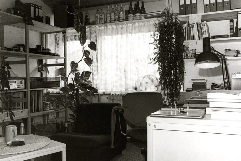 Schwarz-Weiß-Foto: Blick in ein Studentenzimmer in den 70er Jahren. Rechts ein Schreibtisch, links Regale und eine Sitzecke, geradeaus das Fenster, vor dem Gardinen hängen. An den Wänden Regale mit Ordnern. Über dem Fenster ein Regal, auf dem verschiedene Alkoholflaschen stehen.