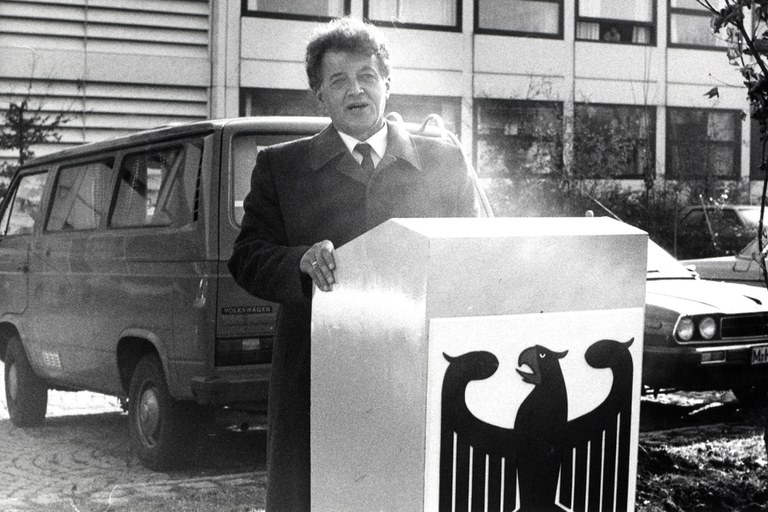 Schwarz-Weiß-Foto: Präsident Wienecke steht an einem Rednerpult, auf dessen Vorderseite das Staatswappen der Bundesrepublik Deutschland, der Adler, abgebildet ist. Der Präsident spricht. Er steht im Freien, hinter ihm parken Autos, im Hintergrund ein Hochschulgebäude.