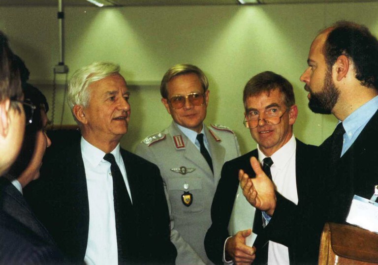 Farbfoto: Bundespräsident Richard von Weizsäcker im Gespräch mit einer Person am rechten Bildrand, vier weitere Personen sind ebenfalls (zum Teil verdeckt) abgebildet, zum Teil in Uniform, zum Teil in Anzug.