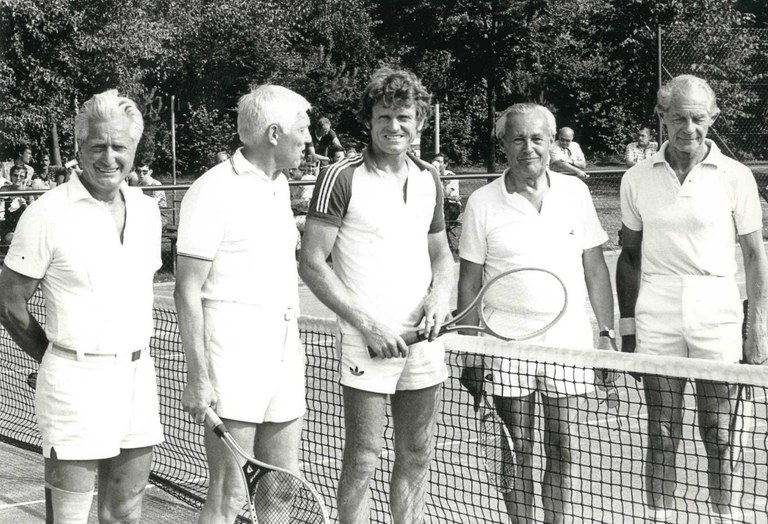 Schwarz-Weiß-Bild: Fünf Männer in Tennisoutfit stehen auf dem Tennisplatz neben dem Netz, drei links im Bild, zwei rechts. In der Mitte mit Tennisschläger in der Hand steht Sepp Maier.