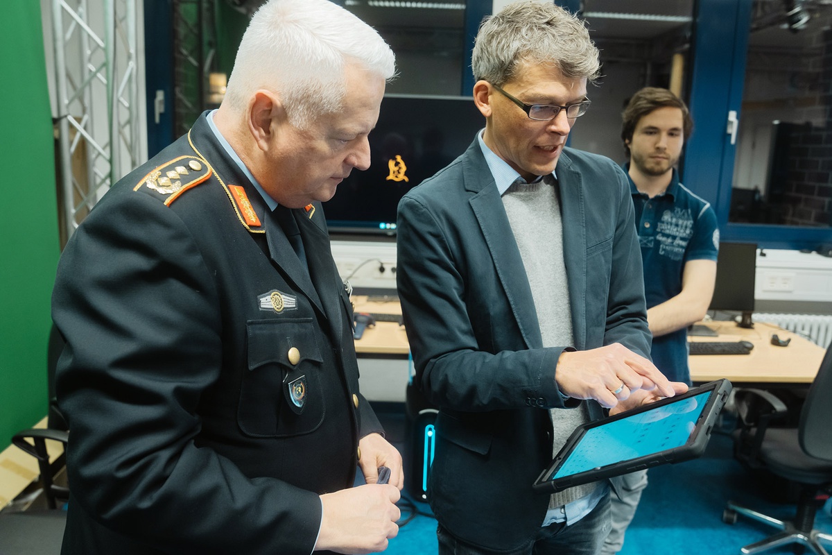 Links im Bild der Generalleutnant in Uniform, rechts neben ihm steht ein Professor von CODE, der ihm etwas auf einem Tablet erklärt, auf das beiden blicken. 