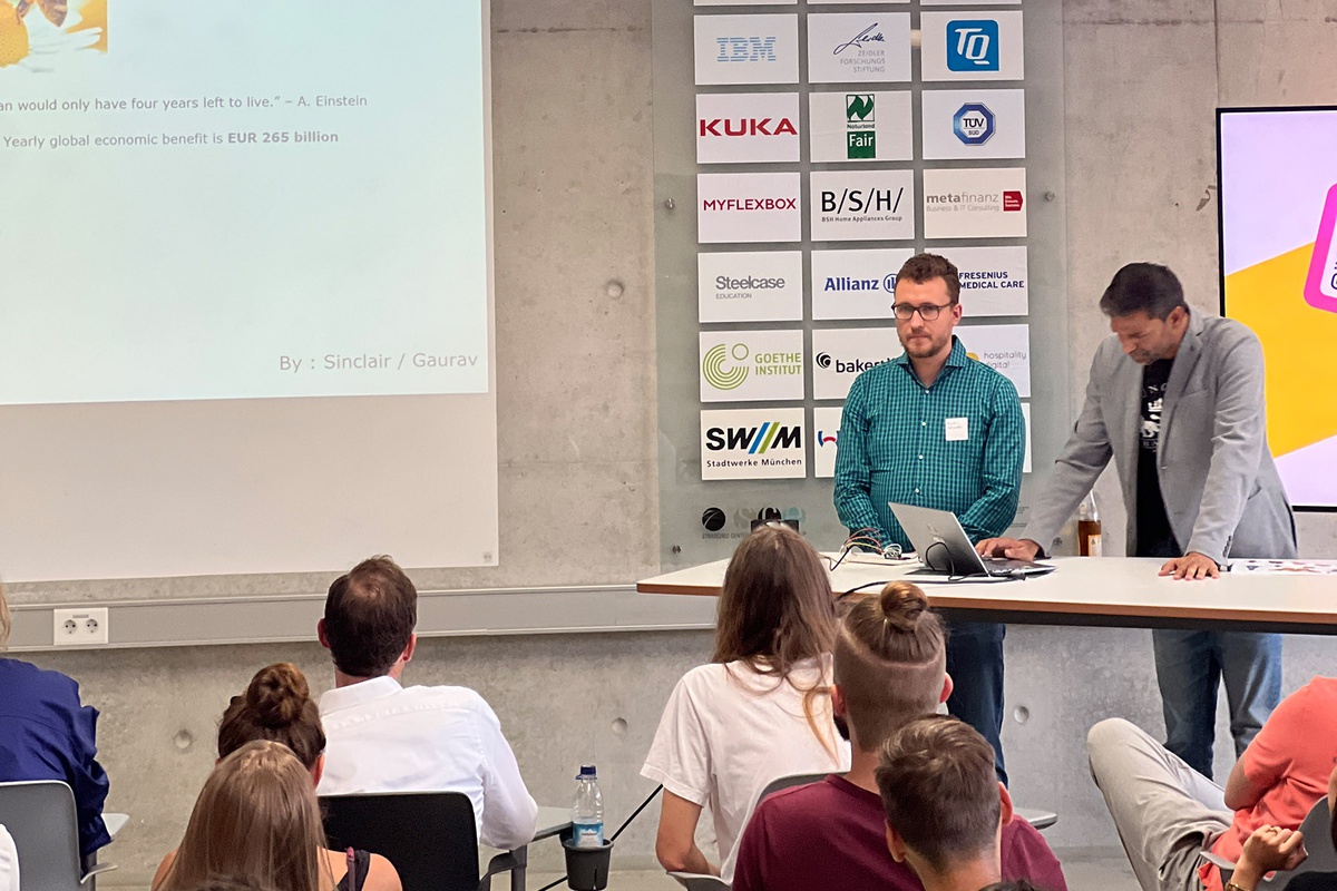 Das Publikum ist von hinten zu sehen, eine Präsentation wird per Beamer an die Wand geworfen, das Foto zeigt jedoch nur Satzfragmente. Rechts von der Leinwand stehen an einem Stehpult zwei junge Männer, einer blickt ins Publikum, der andere bedient den vor ihm stehenden Laptop.