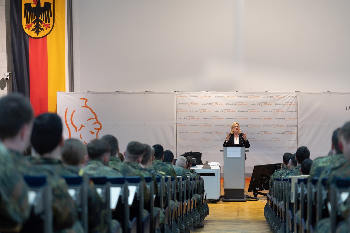 Von hinten den Gang hinunter nach vorne fotografiert: Die Wehrbeauftragte Eva Högl steht im Audimax vorne am Rednerpult und hält einen Impulsvortrag, links und rechts vom Gang in den Sitzreihen sieht man Studierende in Uniform von hinten
