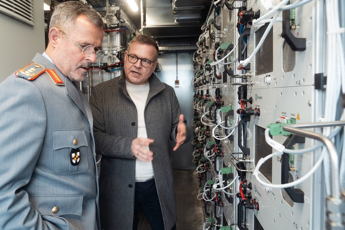 Links im Bild Generalleutnant Nultsch, der auf eine Apparatur mit vielen Kabeln schaut, die sich rechts im Bild befindet. Er hört Prof. Trapp zu, der neben ihm steht und etwas erklärt.