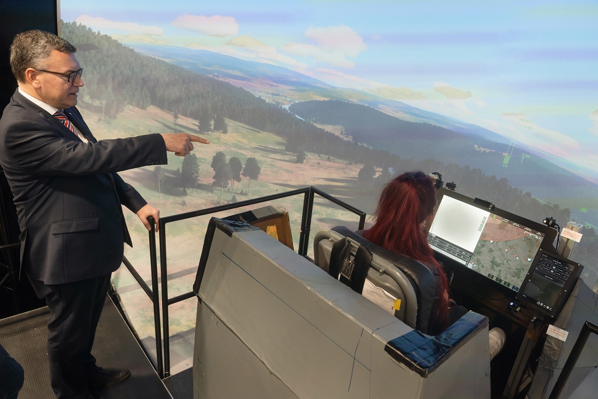 Links im Bild Staatsminister Herrmann, der auf den Bildschirm des Flugsimulators rechts zeigt, in dem eine junge Frau von hinten zu sehen im Cockpit sitzt