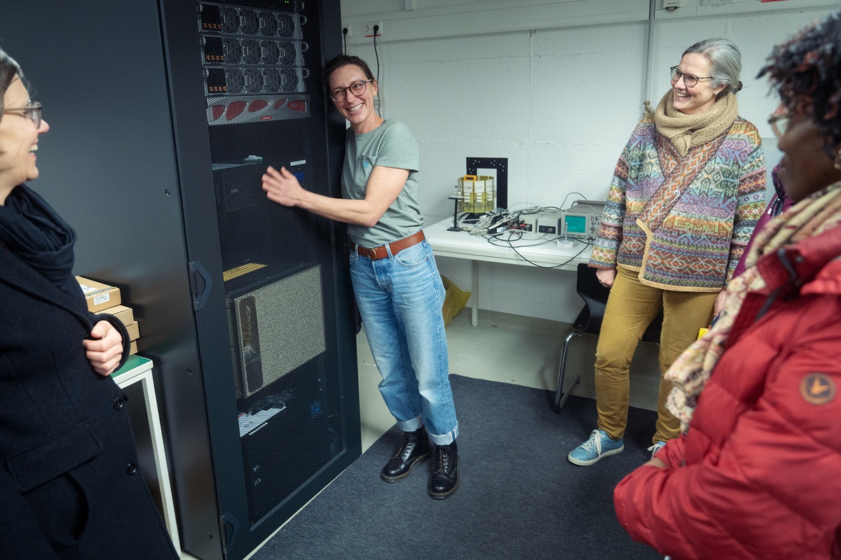 In einem Raum lehnt Prof Riedl sich mit glücklichem Gesichtsausdruck gegen etwas, das wie ein Serverschrank aussieht. Drei Frauen sind mit im Bild und blicken lachend zu Prof. Riedl.