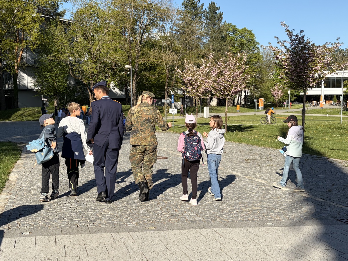 Zwei Studierende beantworten Kinderuni Teilnehmenden Fragen Eine Gruppe läuft in einer Reihe. In der Mitte sind ein Student und eine Studentin in Uniform. Links und rechts sind Kinder. Sie sind in ein Gespräch vertieft.