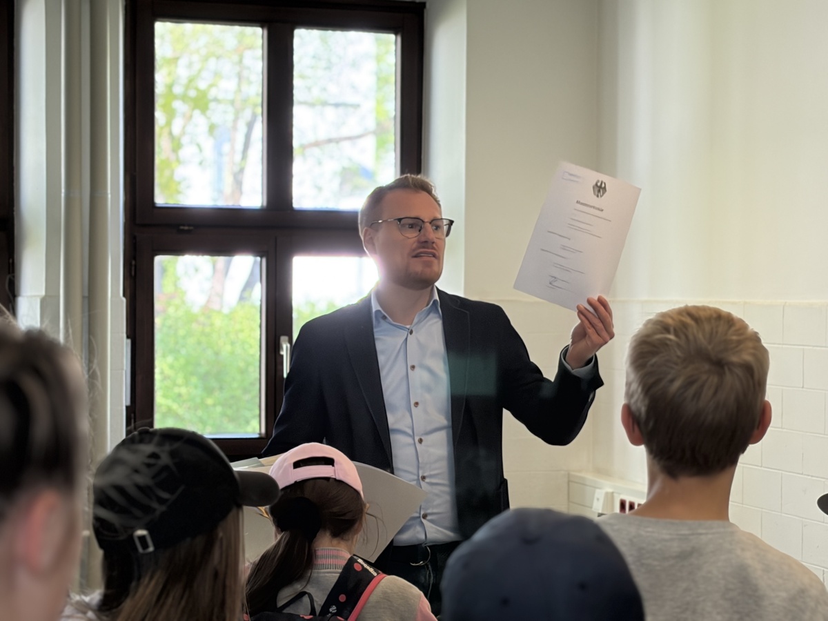 Die Kinderuni Teilnehmenden im Prüfungsamt der UniBw M Im Vordergrund stehen die Schülerinnen und Schüler. Herr Weber vom Prüfungsamt hält eine Urkunde hoch und erklärt, was das Prüfungsamt damit macht.