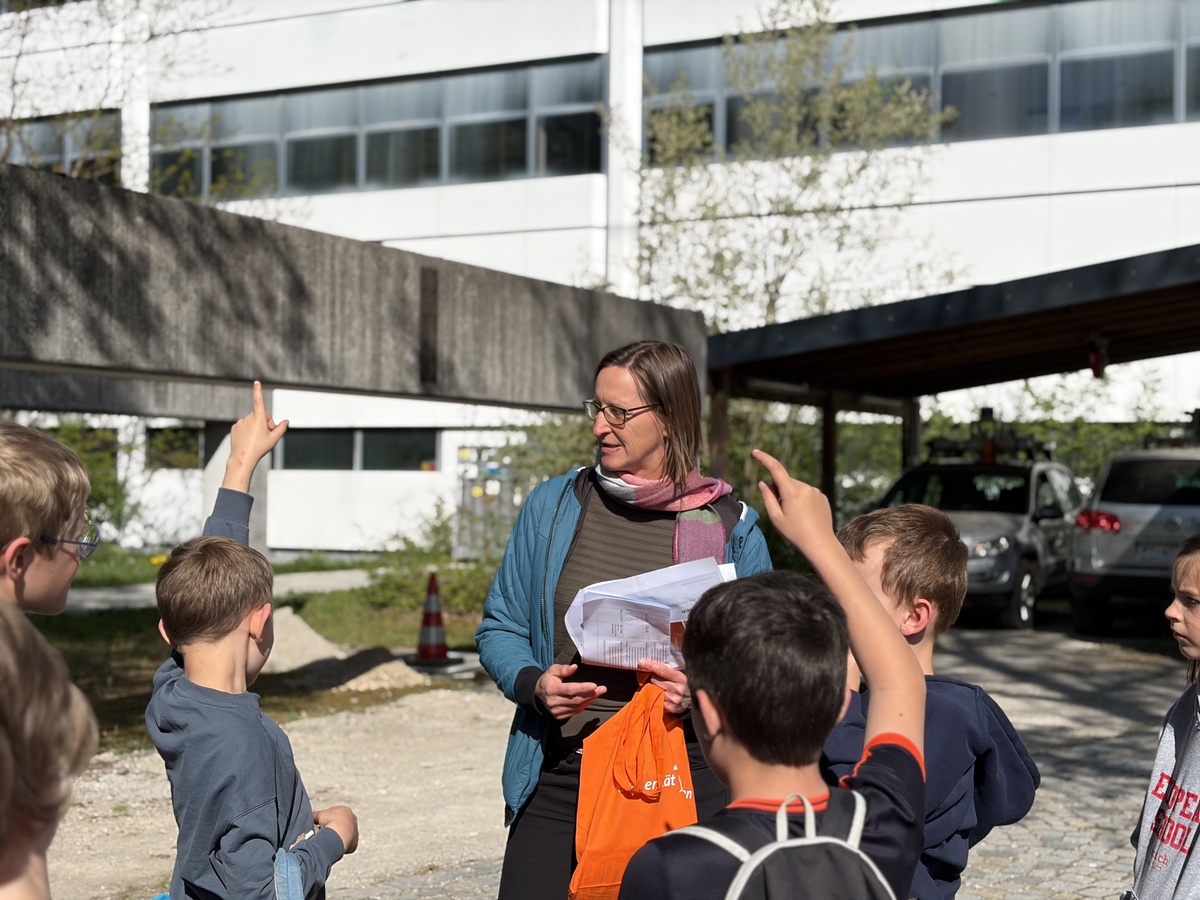 Fragerunde bei Kinderuni auf dem Campus der UniBw M Im Vordergrund sieht man merhere Kinder, die sich melden. Eine Mitarbeiterin der UniBW M sieht ein Kind an und hört sich dessen Frage an.