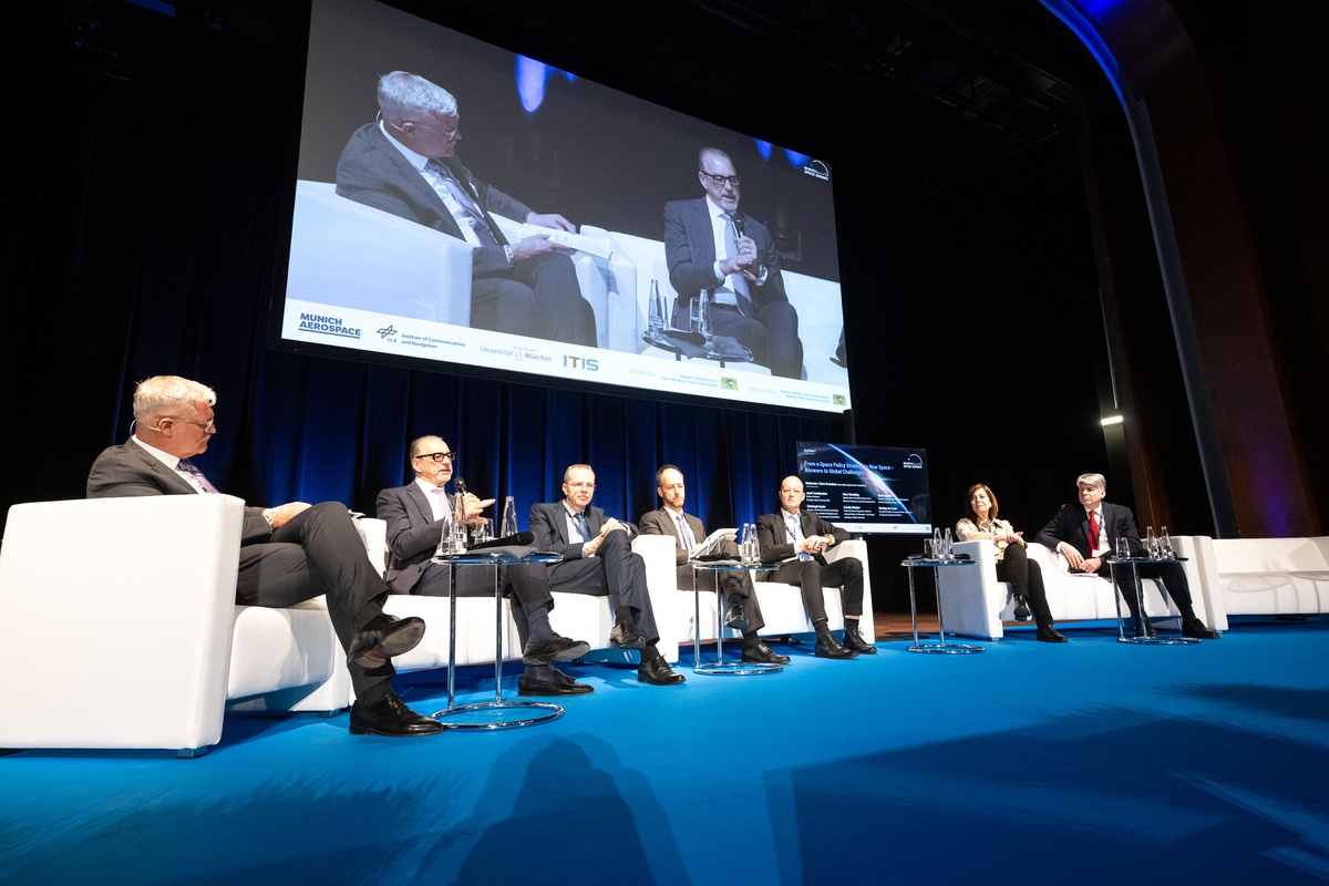 Paneldiskussion zur Eröffnung des Munich Space Summit Sieben PErsonen sitzen auf weißen Sofas und sind einander zugewendet. ein Herr hat ein Mikro in der Hand und spricht.