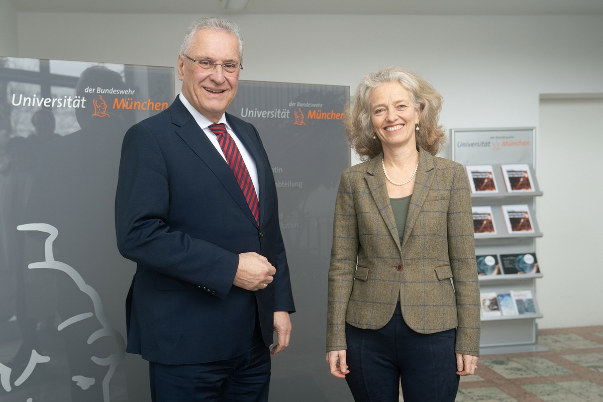 Begrüßung durch Präsidentin Kern Links im Bild innenminister Herrmann, rechts im Bild Präsidentin Prof. Kern. Die beiden stehen im Foyer vor einem grauen Hitnergrund mit dem Universitätslogo und der Athene.