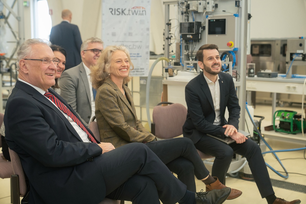 Projekt RISK.twin In einem Raum, in dessen Hintergrund der technische Gerätschaften zu sehen sind, befinden sich links im Bild mehrere Stühle, auf denen unter anderem links im Bild Innenminister Herrmann sitzt, danaben Prof. Kern und rechts Prof. Popp. Alle haben den Blick nach rechts gewandt und schauen auf etwas, das sich außerhalb des Bildes befindet.