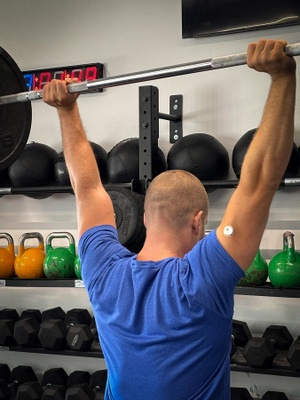 Sportler mit Glukosesensor am Oberarm Rückansicht eines Sportlers, der gerade ein Gewicht in die Höhe streckt. Auf der Rückseite seines rechten Oberarms ist ein keiner, weißer Sensor zu erkennen, der etwa so groß ist wie ein 5-Cent-Stück.
