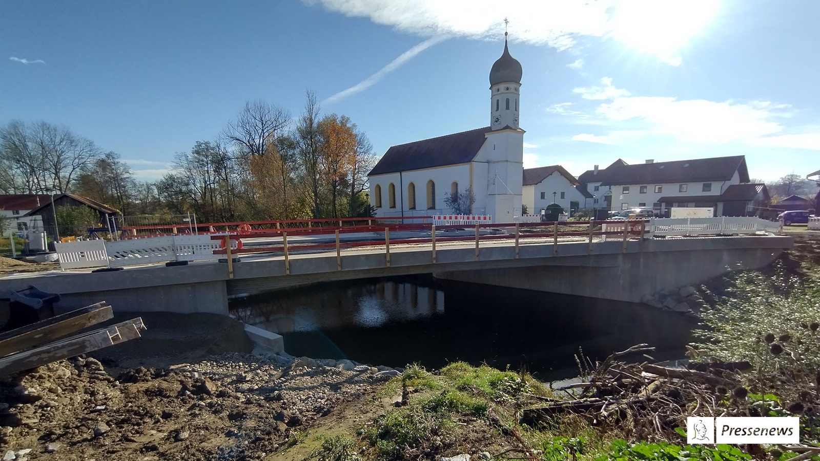 Universität der Bundeswehr und NetApp arbeiten enger zusammen In Schwindegg: Bauarbeiten, eine Brücke mit provisorischem Geländer führt über einen Fluss, an der Uferböschung liegen Stahlträger auf aufgewühlter Erde, daneben aufgeschichtete Äste/Wurzeln, Im Hintergrund auf der anderen Uferseite eine Kirche und Häuser, auch dort Baustelle