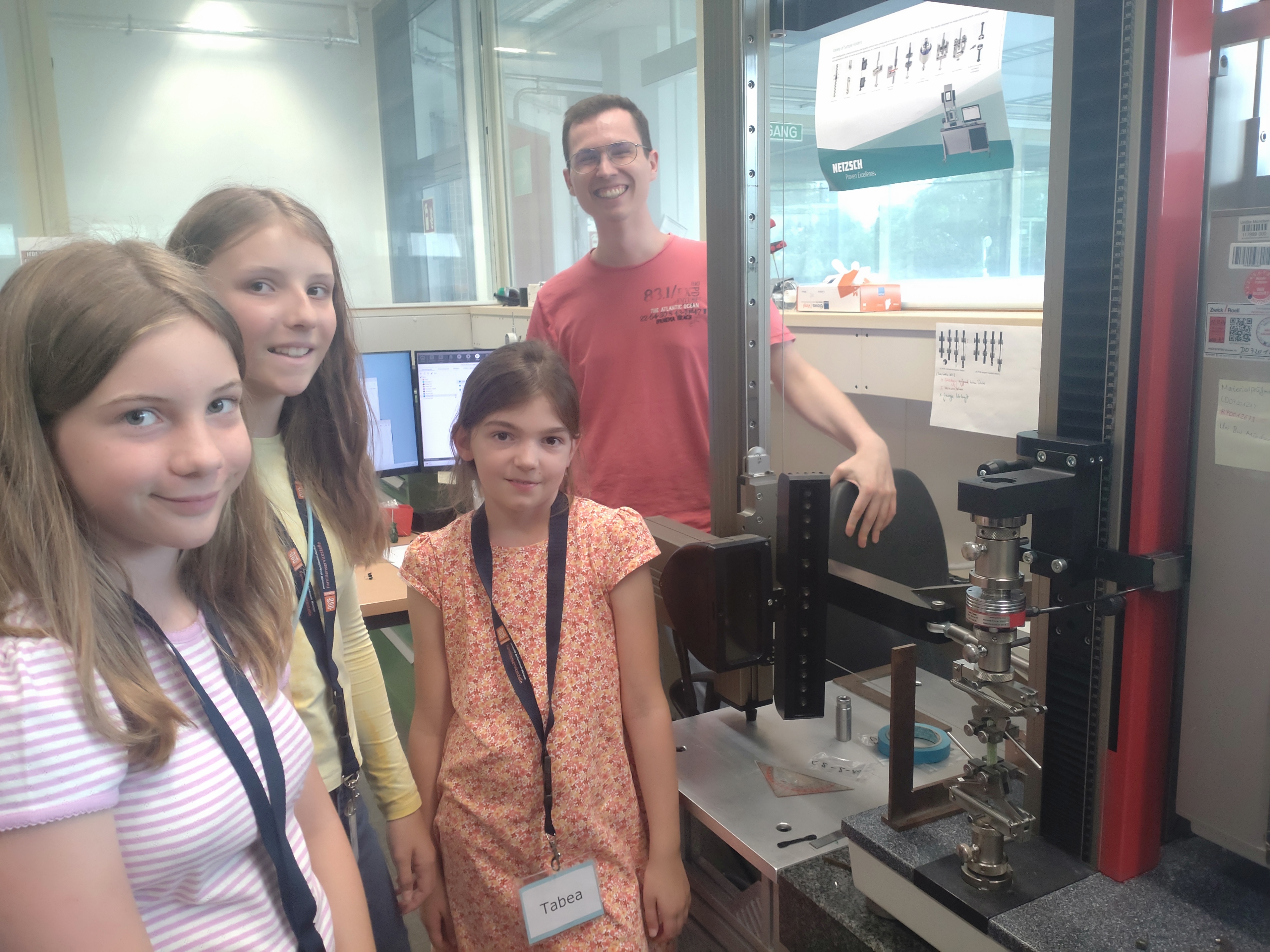 Ein Mitarbeiter zeigt drei Kursteilnehmerinnen Zugversuche mit Süßigkeiten an der Universalprüfmaschine "Zwicky"